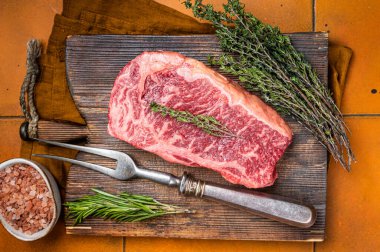 Raw Wagyu striploin, club steak on wooden board with thyme and rosemary ready for cooking. Orange background. Top view.