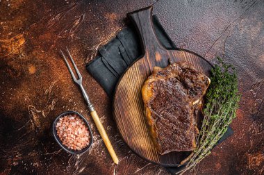 Roast Striploin beef meat steak or club steak on a wooden board with herbs. Dark background. Top view.