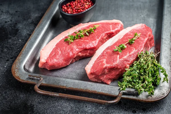 Top sirloin steak, raw beef meat steak with thyme and pepper on steel tray. Black background. Top view.