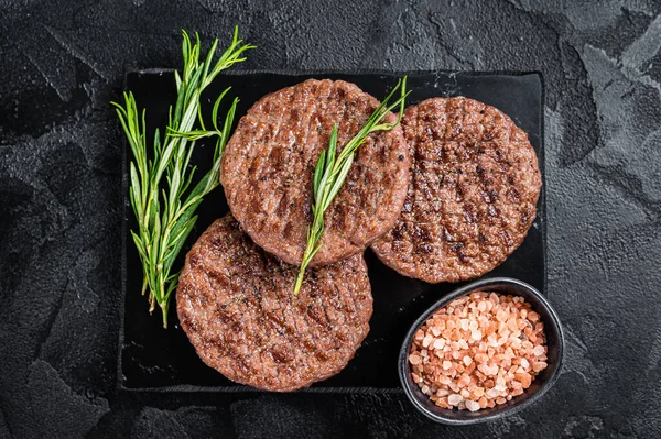 Beef burger patty cutlet for hamburger grilled on BBQ on marble board with rosemary. Black background. Top view.