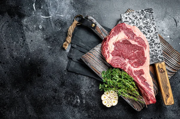 Dry aged Tomahawk rib eye steak, raw beef meat on butcher table. Black background. Top view. Copy space.