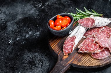 Spanish tapa Fuet sausage slices on wooden cutting board with herbs. Black background. Top view. Copy space.