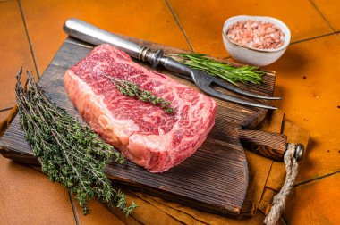 Raw Wagyu striploin, club steak on wooden board with thyme and rosemary ready for cooking. Orange background. Top view.