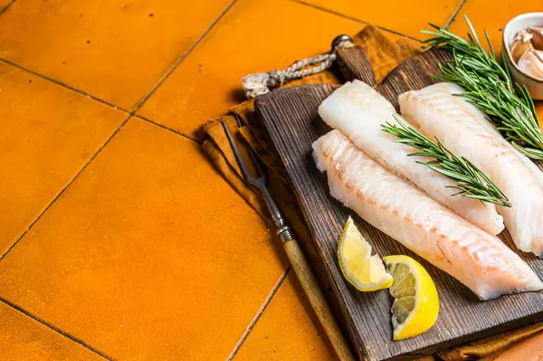 Raw cod fish fillets, codfish with rosemary on wooden board. Orange background. Top view. Copy space.