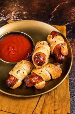 Rolled hot dog sausages baked in puff pastry with ketchup. Dark background. Top view.