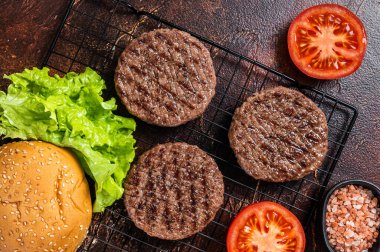 Grilled hamburger patties with tomatoes and seasonings on kitchen table. Dark background. Top view.