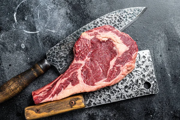 Dry aged Tomahawk rib eye steak, raw beef meat on butcher table. Black background. Top view.
