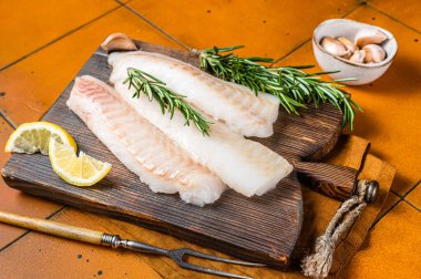 Raw cod fish fillets, codfish with rosemary on wooden board. Orange background. Top view.