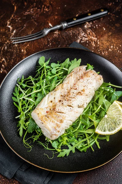 Grilled cod fish fillet served with green salad in a plate. Dark background. Top view.