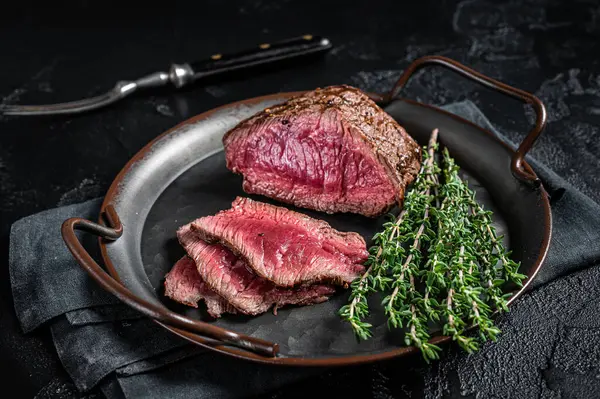 Grilled Rump sirloin steak sliced on a tray with herbs. Black background. Top view.