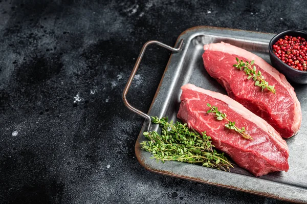 Top sirloin steak, raw beef meat steak with thyme and pepper on steel tray. Black background. Top view. Copy space.