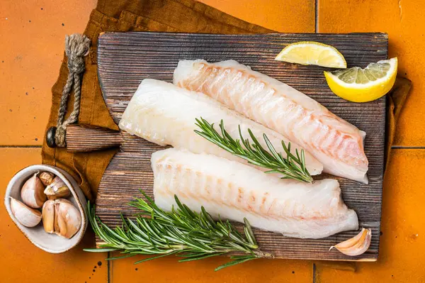 Raw cod fish fillets, codfish with rosemary on wooden board. Orange background. Top view.