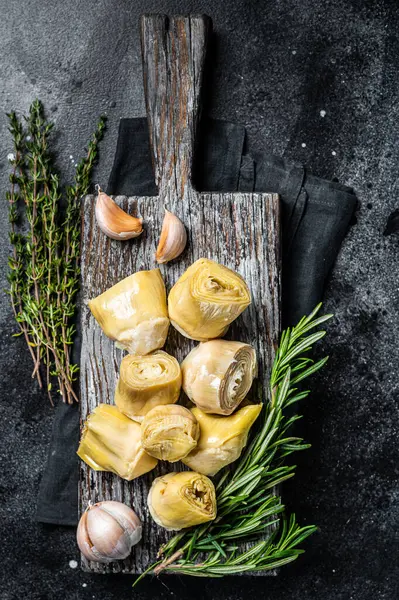 Artichokes hearts marinated with olive oil and herbs, pickled artichoke with garlic on wooden board. Black background. Top view.