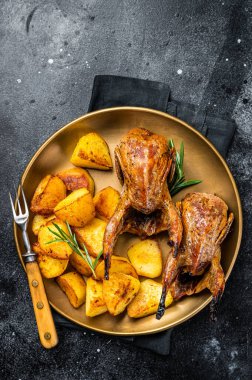 Delicious roasted quails with herbs in a plate with fried potato. Black background. Top view.