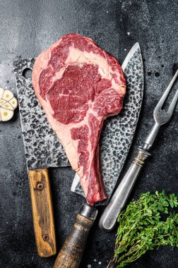 Dry aged raw tomahawk or cowboy beef steak on a black table with knife, cleaver, spice and herbs. Black background. Top view.