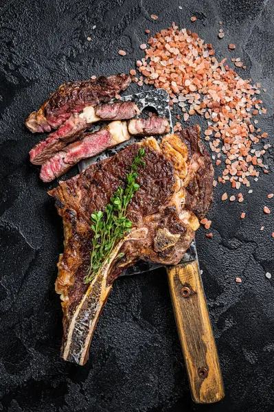 Dry aged grilled Tomahawk or cowboy beef meat steak on a meat cleaver. Black background. Top view.