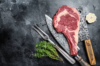 Dry aged raw tomahawk or cowboy beef steak on a black table with knife, cleaver, spice and herbs. Black background. Top view. Copy space.