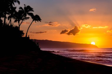 Sun setting behind the mountain over waves and palm trees on Sunset Beach, Hawaii