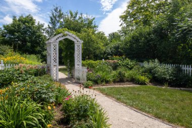 Bitkiler ve çiçeklerle çevrili beyaz boyalı ahşap bir arbala giden yol.