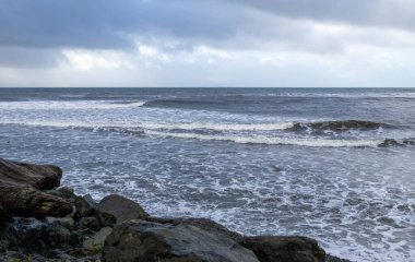 Okyanus dalgaları ve ön plandaki kayalar bulutlu.