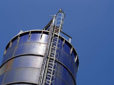 Açık mavi gökyüzüne doğru uzanan metal bir merdiveni olan uzun, mavi bir sanayi silosunun çarpıcı alçak açılı görüntüsü..