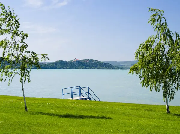 Balaton Gölü 'ndeki pitoresk, serbest bir plajda yeşil çimenler, zarif ağaçlar ve mavi su merdivenleri bulunur. Uzak Tihany Yarımadası' nda, berrak bir yaz gökyüzünün altında ikonik Tihany Manastırı görülebilir..