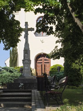 The entrance of the Zamardi Roman Catholic Church, featuring a prominent stone cross from 1862 in the foreground, flanked by benches and lush green bushes, framed by trees under a clear sky.