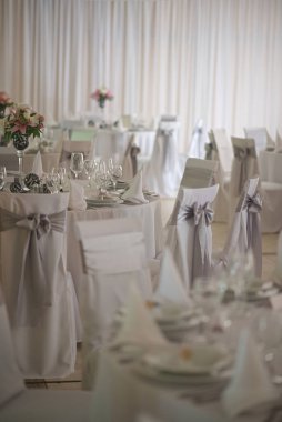 Beyaz masa örtüleriyle süslenmiş masalar, gri yay ile örtülmüş sandalyeler ve yumuşak ışıklandırmanın altında çiçek süslemeleri olan bir etkinlik için hazırlanmış zarif bir karşılama salonu..