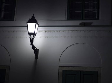 Eski moda bir sokak lambası beyaz bir bina duvarına bağlanmış, gecenin karanlığında parlayan..