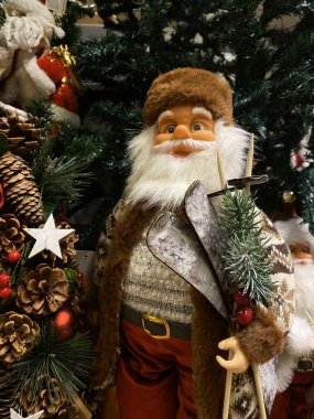 Fotoğrafta, Noel Baba 'nın samimi kış kıyafetleri giymiş bir Noel Baba figürünün olduğu bir festival sahnesi yer alıyor. Noel Baba kahverengi kürklü bir ceket, desenli bir kazak ve altın kemer tokalı kırmızı bir pantolon giyiyor. Elinde bir çift kayak ve sk var.