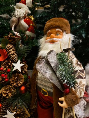 Fotoğrafta, Noel Baba 'nın samimi kış kıyafetleri giymiş bir Noel Baba figürünün olduğu bir festival sahnesi yer alıyor. Noel Baba kahverengi kürklü bir ceket, desenli bir kazak ve altın kemer tokalı kırmızı bir pantolon giyiyor. Elinde bir çift kayak ve sk var.