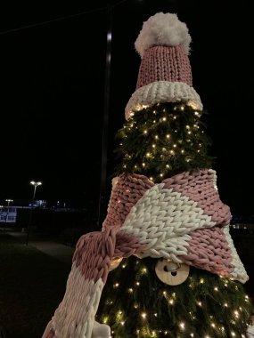 The photo features a Christmas tree decorated in an unusual way. The top of the tree is adorned with a large pink hat with a white pompom, and a massive pink-and-white scarf is wrapped around the tree. The tree is also decorated with large decorative clipart