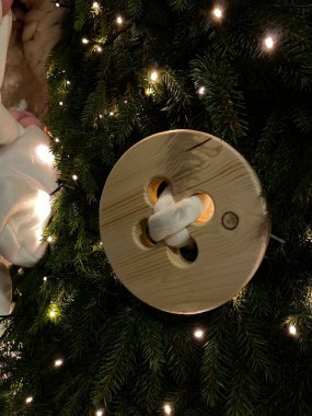 Fotoğrafta, büyük bir ahşap düğme süsüsüyle süslenmiş bir Noel ağacının yakın çekimi yer alıyor. Düğme dört delikli daireseldir, beyaz bir kumaşın geçirildiği ve düğümlendiği yerdir. Ağaç da küçük, sıcak beyaz iplikçiklerle süslenmiş.