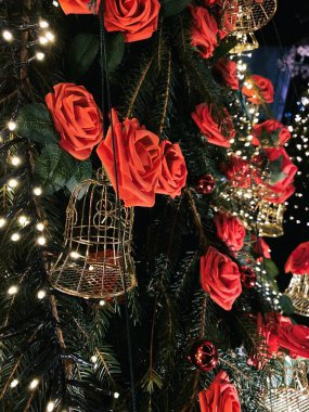 Fotoğrafta, kırmızı güller ve sayısız küçük beyaz ışıkla aydınlatılmış küçük altın kuş kafesleriyle süslenmiş bir Noel ağacı yer alıyor. Arka planda, beyaz ışıklara sarılmış başka bir ağaç festival atmosferine katkı sağlıyor. Kombinasyon: 