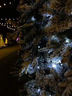 Beyaz ışıklarla süslenmiş ve yapay karla kaplı bir Noel ağacının fotoğrafı. Ağaç, arka planda diğer festival ışıkları ve dekorasyonlarla birlikte açık havada yer almaktadır. Karla kaplı dallar, kış harikalar diyarı etkisi yaratır..