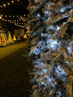 A photograph of a Christmas tree adorned with white lights and covered in artificial snow. The tree is located outdoors, with other festive lights and decorations visible in the background. The snow-covered branches create a winter wonderland effect. clipart