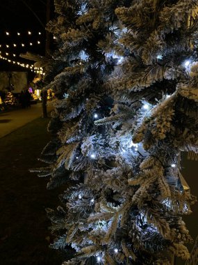 Beyaz ışıklarla süslenmiş ve yapay karla kaplı bir Noel ağacının fotoğrafı. Ağaç, arka planda diğer festival ışıkları ve dekorasyonlarla birlikte açık havada yer almaktadır. Karla kaplı dallar, kış harikalar diyarı etkisi yaratır..