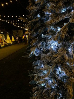 Beyaz ışıklarla süslenmiş ve yapay karla kaplı bir Noel ağacının fotoğrafı. Ağaç, arka planda diğer festival ışıkları ve dekorasyonlarla birlikte açık havada yer almaktadır. Karla kaplı dallar, kış harikalar diyarı etkisi yaratır..