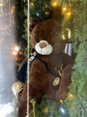 A photograph of a large teddy bear sitting within a green Christmas wreath adorned with small lights and hanging ornaments. The teddy bear is wearing a dark blue sash with the text 