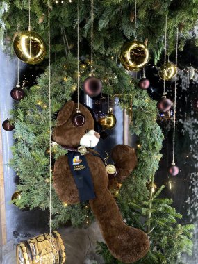 A photograph of a large teddy bear sitting within a green Christmas wreath adorned with small lights and hanging ornaments. The teddy bear is wearing a dark blue sash with the text 