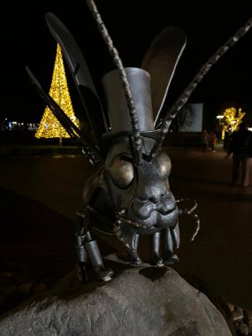 The photo features a detailed metal sculpture of an insect wearing a top hat, positioned on a rock. The insect has prominent eyes, antennae, and legs made of metal parts. In the background, a large illuminated Christmas tree made of lights indicates  clipart