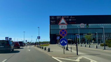 Alışveriş merkezi ve park alanı yakınında trafik ışıkları ve yol işaretleri olan şehir yolu, şehir ulaşımı ve kavşak manzarası. Yüksek kalite fotoğraf. Kaunas, Kauno m. SAv., Litvanya 04.27.2025