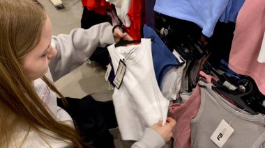 Moda bir giyim mağazasında kot pantolon almak için kapüşon giyen genç bir kadın. Moda alışverişi, modern yaşam tarzı, gündelik moda, ergen modası, perakende deneyimi. Bir Giyim Mağazasında Çok Giysi Askısı Tutan Kız, Alışveriş Konsepti, Fa