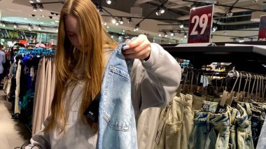 Moda bir giyim mağazasında kot pantolon almak için kapüşon giyen genç bir kadın. Moda alışverişi, modern yaşam tarzı, gündelik moda, ergen modası, perakende deneyimi. Bir Giyim Mağazasında Çok Giysi Askısı Tutan Kız, Alışveriş Konsepti, Fa