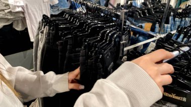 Moda bir giyim mağazasında kot pantolon almak için kapüşon giyen genç bir kadın. Moda alışverişi, modern yaşam tarzı, gündelik moda, ergen modası, perakende deneyimi. Bir Giyim Mağazasında Çok Giysi Askısı Tutan Kız, Alışveriş Konsepti, Fa