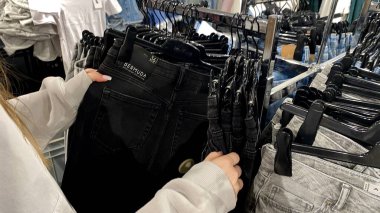 Moda bir giyim mağazasında kot pantolon almak için kapüşon giyen genç bir kadın. Moda alışverişi, modern yaşam tarzı, gündelik moda, ergen modası, perakende deneyimi. Bir Giyim Mağazasında Çok Giysi Askısı Tutan Kız, Alışveriş Konsepti, Fa