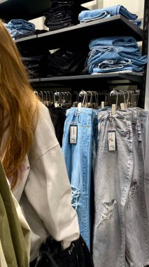 Moda bir giyim mağazasında kot pantolon almak için kapüşon giyen genç bir kadın. Moda alışverişi, modern yaşam tarzı, gündelik moda, ergen modası, perakende deneyimi. Bir Giyim Mağazasında Çok Giysi Askısı Tutan Kız, Alışveriş Konsepti, Fa