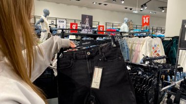 Moda bir giyim mağazasında kot pantolon almak için kapüşon giyen genç bir kadın. Moda alışverişi, modern yaşam tarzı, gündelik moda, ergen modası, perakende deneyimi. Bir Giyim Mağazasında Çok Giysi Askısı Tutan Kız, Alışveriş Konsepti, Fa