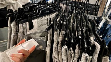 Moda bir giyim mağazasında kot pantolon almak için kapüşon giyen genç bir kadın. Moda alışverişi, modern yaşam tarzı, gündelik moda, ergen modası, perakende deneyimi. Bir Giyim Mağazasında Çok Giysi Askısı Tutan Kız, Alışveriş Konsepti, Fa