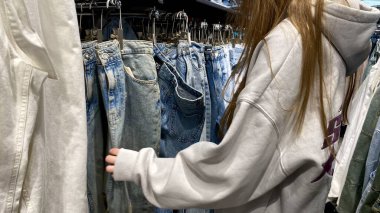 Moda bir giyim mağazasında kot pantolon almak için kapüşon giyen genç bir kadın. Moda alışverişi, modern yaşam tarzı, gündelik moda, ergen modası, perakende deneyimi. Bir Giyim Mağazasında Çok Giysi Askısı Tutan Kız, Alışveriş Konsepti, Fa
